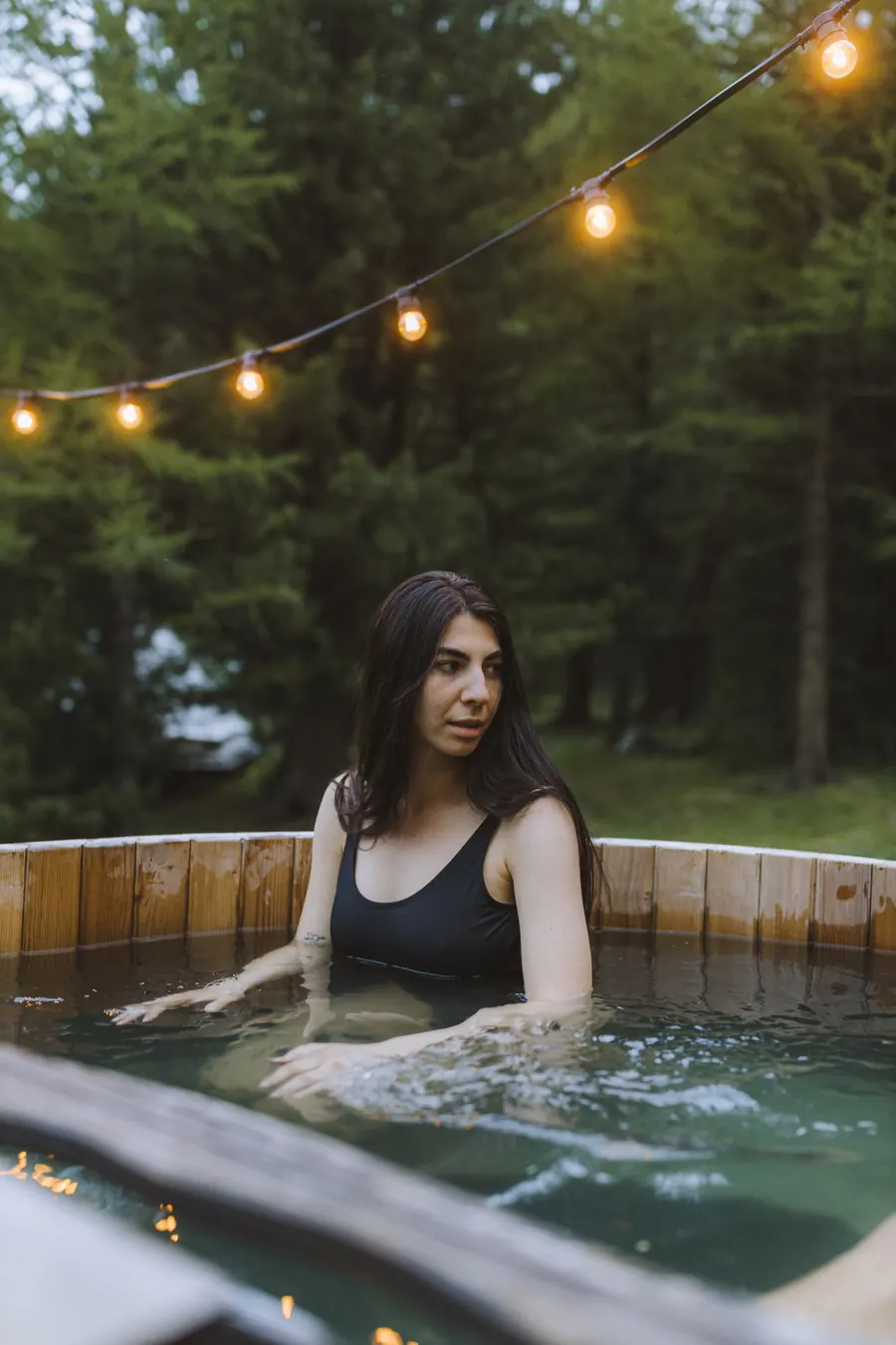 A woman in a hot tub under string lights at dusk
